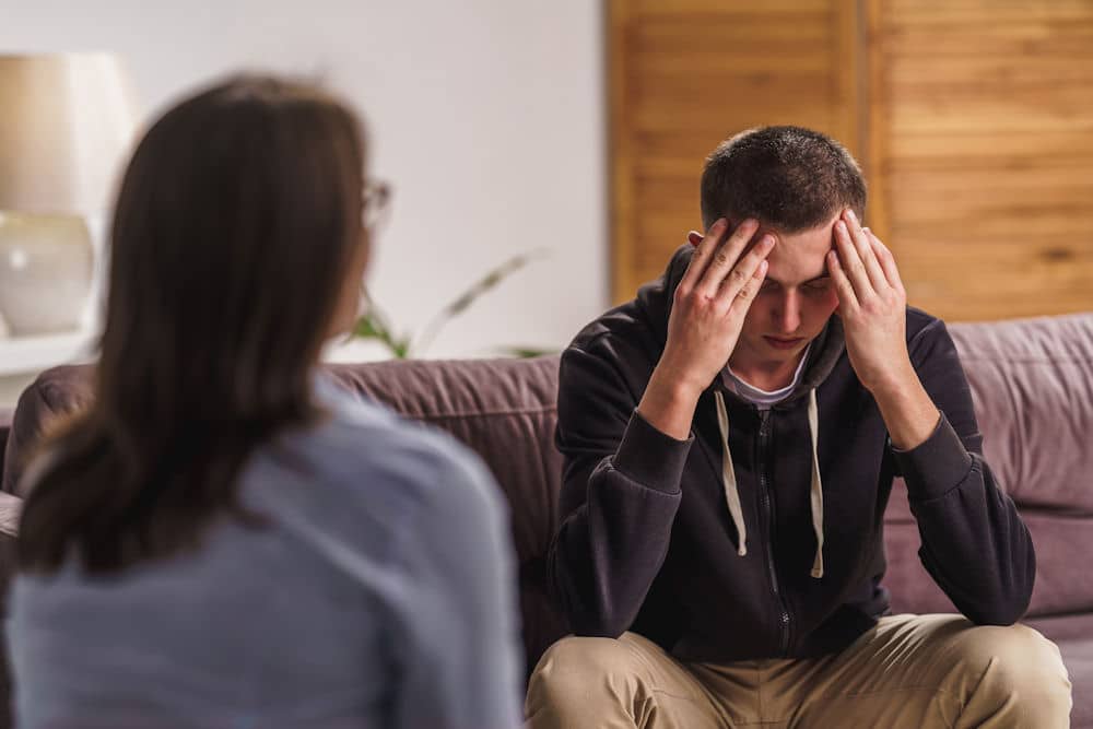 Frustrated young man in hoodie holding head while talking to unseen woman in glasses