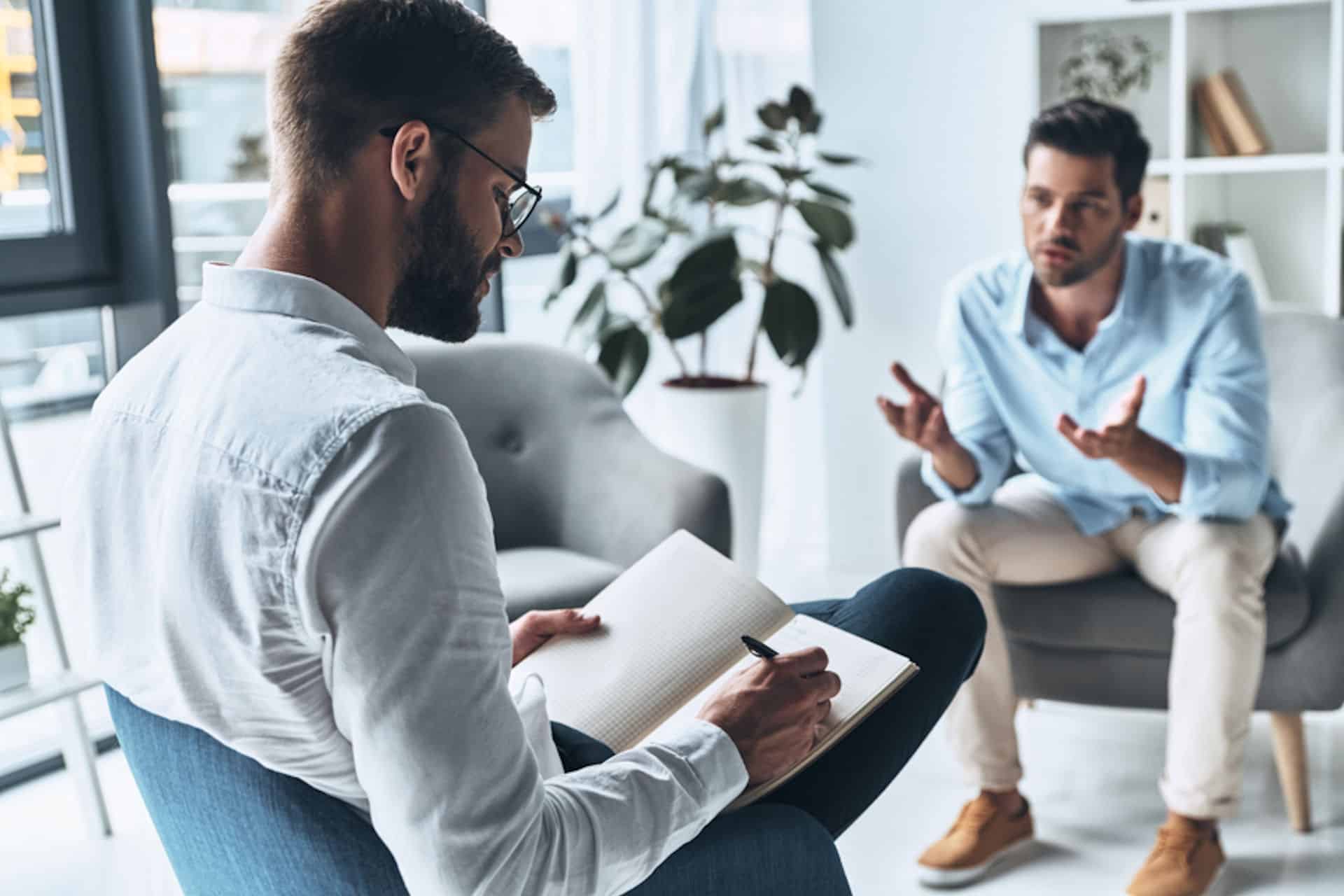 Seated man describing depression to second man taking notes in notebook