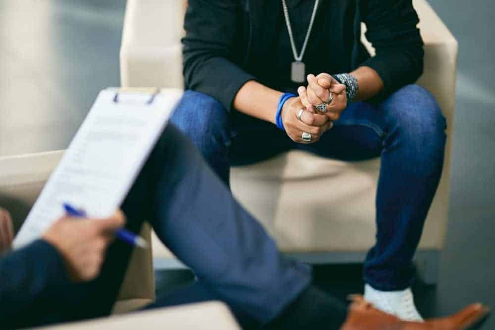 Chest-down view of seated man wearing many rings speaking to therapist with clipboard