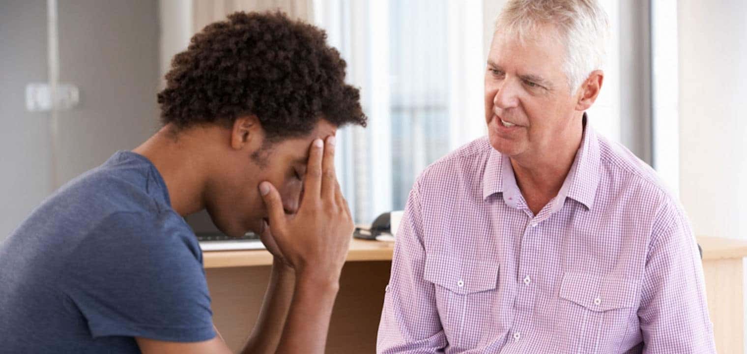 Young man holds head while older man takes notes on notepad