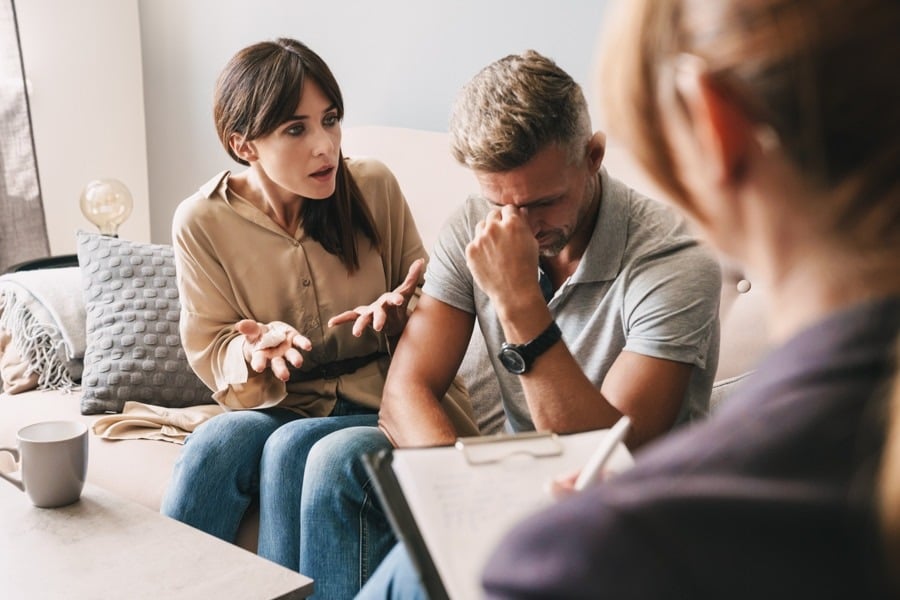 Woman argues with frustrated man sitting next to her on couch while therapist holding clipboard looks on